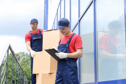 Two male removal workers dressed in red T-shirts, blue overalls, and navy caps are outdoors beside a modern glass-fronted building on a residential street. The worker in the foreground is holding a clipboard and wearing white gloves, appearing to record information, while the other is standing behind, carrying several cardboard boxes stacked on top of each other. The boxes are plain brown cardboard with visible tape along the edges, and the workers are positioned near a metal railing on their left. The building features large reflective glass panels, reflecting parts of the environment, and the sky above shows cloudy weather. The scene suggests a house or apartment removals operation in a calm, suburban area, potentially within the Tufnell Park or nearby postcode region, and demonstrates careful handling and record-keeping during a relocation process. The natural lighting highlights the textures of the cardboard boxes and the workers’ uniforms, emphasizing a professional and organized move aligned with house removal services.