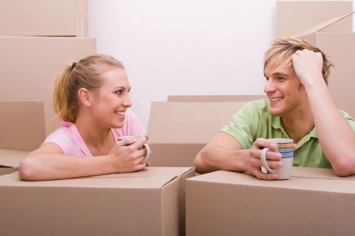 A young woman and a young man with light blond hair and fair skin are sitting inside a room filled with cardboard moving boxes, which suggests a house relocation or packing process. The woman, wearing a light pink shirt, is smiling and holding a smartphone in her hands, while the man, dressed in a light green T-shirt, is smiling back and resting his head on his left hand, holding a striped ceramic mug in his right hand. They are positioned behind stacked cardboard boxes on a plain, off-white wall background, indicating an indoor environment possibly within a residential property in Tufnell Park or nearby London. The boxes are closed and arranged in a casual manner, with some stacked on top of each other, implying an ongoing move or packing activity typical of local house removals. The lighting is even and soft, creating a warm, relaxed atmosphere suitable for a domestic house removals service in North London areas like Tufnell Park.