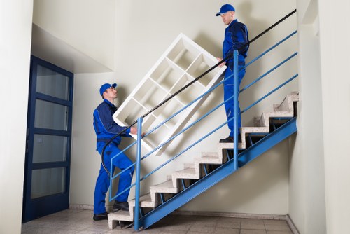 Two professional removal workers from Tufnell Park Man with Van are in a residential staircase area, engaged in lifting and carrying a large white shelving unit with multiple compartments made of wood or composite material. The worker on the lower level, dressed in blue work clothing and a cap, supports the unit from below, while the taller worker, also in blue attire and a cap, is positioned on the staircase's upper landing, guiding the furniture up the stairs. The staircase has a modern design with metal railings painted in a matching blue colour, and steps covered with dark, textured material. The surrounding interior features white walls and a large window to the left, allowing natural light to illuminate the scene. The environment indicates a clean, well-maintained residential property, suitable for house removals or furniture relocations, with the scene reflecting careful handling of household items as part of the moving process in the Tufnell Park area.