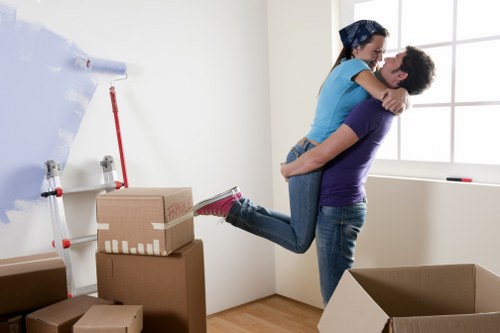 In a bright, minimally decorated room with cream walls and a large window allowing natural light, a man and a woman are engaged in house moving. The man, dressed in a dark purple t-shirt and jeans, is holding the woman, who is wearing a blue top and jeans, off the ground in an embrace. The woman has her arms around the man's neck and appears to be smiling, suggesting a cheerful moment during the removal process. On the floor beside them, there are several packed cardboard boxes, some sealed with packing tape, and a large houseplant is partially visible in the background. The scene includes a white wall-mounted shelf with a red-handled tool and a white roll of paper or packing material, indicating packing activity. The room is clean and well-lit with natural daylight coming through the window, and the overall setting reflects a typical residential space in Tufnell Park, suitable for house removals and relocation services by Tufnell Park Man with Van.