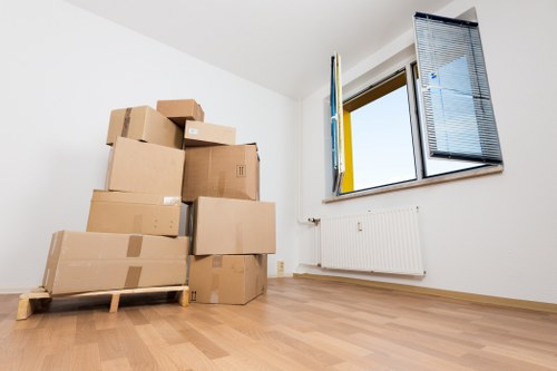 The image depicts a bright, minimally furnished room with white walls and a light wooden floor, likely within a residential property in Tufnell Park. In the foreground, there is a stack of cardboard moving boxes of various sizes, some sealed with packing tape and others open, placed on a wooden pallet for ease of transport. The boxes are arranged in a loose pile near the corner of the room. On the right side of the image, a large window with black framing is partially open, revealing blue sky outside; the window has black external shutters that are folded back, allowing natural daylight to fill the space. Below the window, a white radiator is mounted on the wall. The scene resembles a typical early stage of a house removal process, with household items packed and ready for transport, illustrating a tidy, clean interior environment. This setting is consistent with services provided by Tufnell Park Man with Van within the N19 postcode area, focusing on efficient and professional removals in North London.