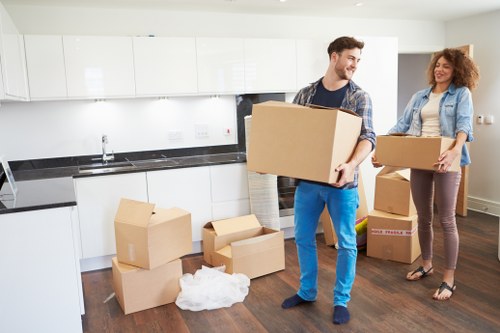 A young man and woman inside a modern kitchen, surrounded by moving boxes of various sizes and materials, including cardboard boxes with tape and some open, revealing packaging paper. The man, with dark hair and wearing a checkered shirt and jeans, is carrying a large cardboard box, looking to his right with a smile. The woman, with curly reddish hair and dressed in a denim jacket and brown trousers, holds a medium-sized box and faces the man, also smiling. The kitchen features white cabinetry, a black countertop, a stainless steel sink, and a white tiled backsplash. The floor is dark wood, creating a warm contrast to the bright, clean environment. The scene appears to be part of a house relocation or home moving process, with the background showing a residential space typical of Tufnell Park and the postcode area associated with North London, indicating a local house removals context. The lighting is natural, suggesting daytime, emphasizing the organized and professional atmosphere of the move orchestrated by Tufnell Park Man with Van, a local removals service provider.