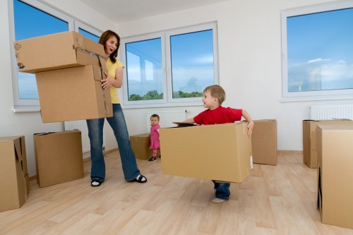 In a bright, spacious room with white walls and large windows showing a blue sky with some clouds, a woman with shoulder-length brown hair, wearing a yellow top and jeans, is holding three large cardboard boxes stacked together near her waist, preparing for a house move. Beside her, a young boy with blonde hair, dressed in a red shirt and jeans, is carrying a smaller cardboard box with both hands, walking across the light wood floor. A small girl in a pink dress stands nearby, observing the activity. Other cardboard boxes of various sizes are scattered throughout the room, some placed on the floor and others stacked against the wall. The room appears to be part of a residential property in Tufnell Park, with natural daylight illuminating the scene, suggesting a calm, organized movescape typical of house removals services offered by Tufnell Park Man with Van.