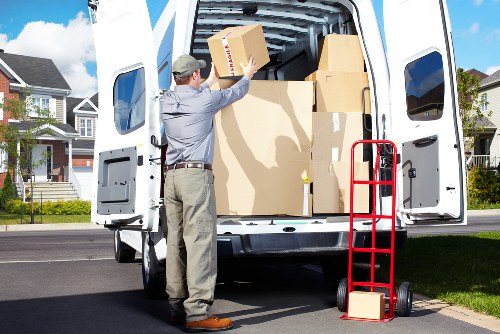 A man wearing a grey shirt, beige trousers, and a cap is lifting a medium-sized cardboard box into the open back of a white removals van parked on a residential street in Tufnell Park, London. The van's interior is filled with various-sized cardboard boxes, some stacked on top of each other, ready for a house relocation. A red hand truck with a small cardboard box resting on its platform is positioned on the pavement next to the van. In the background, there are modern terraced houses with brick facades, front gardens, and well-maintained lawns, indicating a typical London suburb setting. The scene is illuminated by bright daylight, suggesting clear weather, and the precise placement of boxes and equipment reflects an organized approach to property removals, with the focus on safe handling and efficient loading gear for a professional move in North London postcode area.