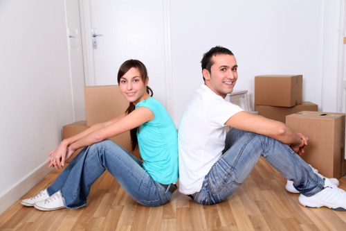 A young man and woman sit back-to-back on a light wooden floor inside a residential property in Tufnell Park, North London, with several cardboard moving boxes stacked around them. The woman, wearing a turquoise T-shirt and blue jeans, is smiling and facing towards the camera, with her legs extended forward and hands resting on her knees. The man, dressed in a white T-shirt and blue jeans, also smiles at the camera, with his legs stretched out and arms relaxed on his knees. The background features a white wall, a white door to the left, and a small white side table next to the boxes, indicating a typical hallway or living space in a house preparing for a removal. The environment appears clean, well-lit with natural light, and uncluttered, suggesting an organized moving process. This scene exemplifies a typical pause during a house relocation, with furniture and personal belongings packed for transportation by Tufnell Park Man with Van, serving clients in the local area and nearby postcode regions.