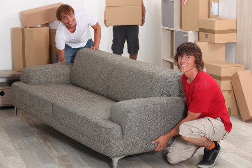 Two young men are in a furnished room that appears to be a part of a residential house, preparing to move a grey fabric upholstered sofa with a textured finish. One man, wearing a white T-shirt and jeans, is crouched behind the sofa, holding its side and looking toward the camera with a slight smile. The other man, dressed in a red T-shirt and beige shorts, is kneeling to the right of the sofa, grasping the front right corner, also smiling at the camera. The room has light wood flooring and a white shelving unit on the right filled with cardboard boxes of various sizes, some open, suggesting they are still packing or unpacking. Additional cardboard boxes are stacked against the wall in the background, along with a hallway or doorway leading to other parts of the house. The lighting is bright and natural, indicating daytime, and the scene conveys a typical house removal activity, with the individuals handling furniture carefully during a relocation process, consistent with services offered by Tufnell Park Man with Van in North London, within the N7 postcode area.