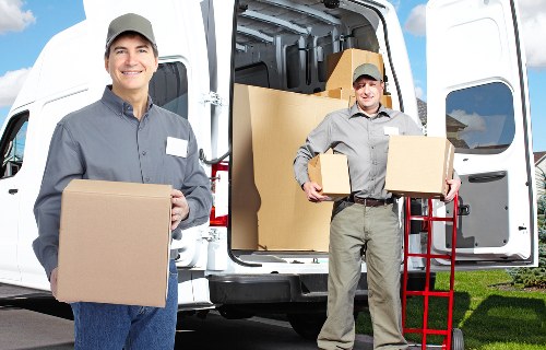 The image shows two professional removals staff members dressed in grey uniforms and caps, standing outdoors next to a white removals van with its rear doors open. The worker on the left is smiling while holding a large cardboard box, and the worker on the right, also smiling, is carrying two smaller cardboard boxes, one in each hand, with a red hand trolley positioned nearby. Visible inside the van are several packed cardboard boxes, some stacked and others positioned upright, ready for transport. The setting is a residential area, possibly in Tufnell Park, with green grass and a clear sky in the background, indicating daytime. The scene captures the process of loading household items for a house removal or relocation service, emphasizing careful handling of packed boxes and professional service from Tufnell Park Man with Van, specialists in removals within the local area.