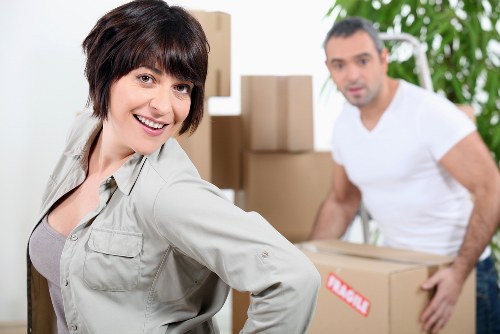 A woman with short dark hair, wearing a light-colored shirt, smiling at the camera while standing in a residential interior filled with cardboard boxes and packing materials, with a man in a white t-shirt in the background carrying a box and a few more boxes stacked nearby. The setting appears to be a house in Tufnell Park, London, during a house removal or moving process, with natural light illuminating the space and some greenery visible through a window or door. The scene highlights domestic moving activity, with household furniture and personal belongings prepared for transport, supporting the scope of a professional removals service focused on eco-friendly and sustainable practices.