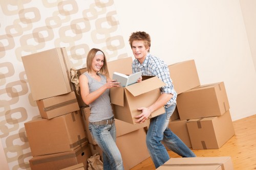 A young man and woman are standing inside a residential property with light-colored walls featuring a subtle geometric pattern, surrounded by stacked cardboard boxes used for house removals. The woman, dressed in a grey sleeveless top and denim jeans, is holding a tablet device and smiling, while the young man, wearing a blue plaid shirt and jeans, is carrying a large cardboard box. Several packed boxes of varying sizes are placed on the wooden floor, suggesting an active packing or moving process. The scene appears to be in a living area or hallway, with natural lighting illuminating the space, and no other furniture or household items visible. The environment reflects a typical house relocation in the UK, with the focus on packing and handling household items, consistent with the services offered by Tufnell Park Man with Van in the postcode area surrounding N7, North London.