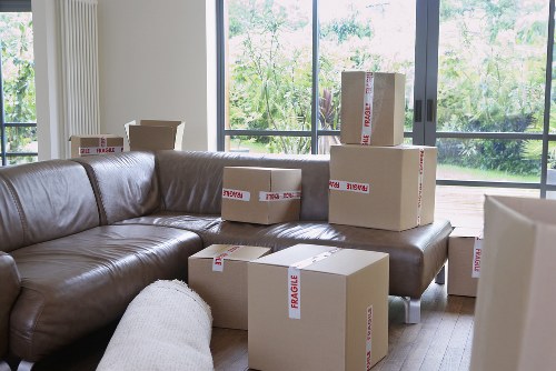 A living room interior showing a brown leather sectional sofa positioned against large floor-to-ceiling windows, revealing a green outdoor garden with trees. Several cardboard moving boxes, some marked with red 'fragile' labels, are stacked on and around the sofa, with a few placed on the wooden floor. The boxes vary in size and are in natural brown cardboard, with packing tape visible on some. The room is well-lit by natural daylight, creating a bright and open environment suitable for house removals in the area. The scene captures the process of packing and preparing for a move, consistent with services offered by Tufnell Park Man with Van, a company specializing in house removals. The setting suggests a residential property in London, with the boxes indicating an ongoing transition or relocation within the local area.
