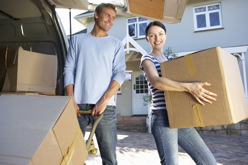 A man and a woman, both smiling, are engaged in a house removal process outdoors on a residential street. The man, dressed in a light blue long-sleeved shirt and dark jeans, is lifting a medium-sized cardboard box with yellow packing tape visible along the edges. He is standing beside an open removals van, which has more cardboard boxes and packing materials visible inside. The woman, wearing a striped short-sleeved top and jeans, is carrying a larger cardboard box, holding it securely with both hands. Behind them, the house features white-framed windows and a front door, with the driveway and a paved area visible in the foreground. The scene is well-lit with natural daylight, emphasizing the practical task of house moving, typical of Tufnell Park area in London, as part of professional removals services offered by Tufnell Park Man with Van.