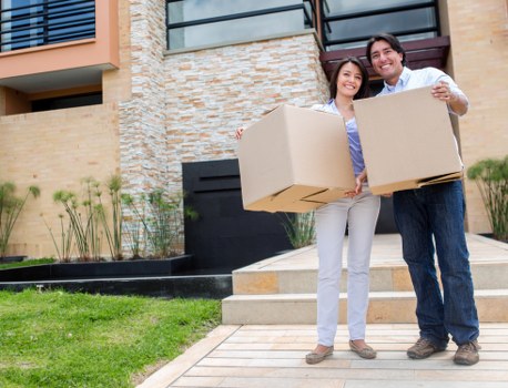 A young couple standing outside a modern residential building with brick and stone exterior, holding large cardboard boxes for moving. The woman, on the left, is wearing light-colored trousers and a casual top, smiling as she holds a box in her right hand and a larger box in her left. The man, on the right, is dressed in dark trousers and a light shirt, also smiling, supporting the boxes with both hands. They are positioned on a paved pathway leading to the house entrance, which has concrete steps and a black door with a small porch area. To the left of the couple, there is a well-maintained green lawn with small garden plants and shrubs in a raised bed bordered by black edging. The scene is set during daylight with natural lighting, capturing a typical house removal or relocation moment, consistent with services offered by Tufnell Park Man with Van in the London postcode area, subtly indicating a residential setting in North London.