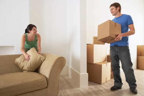 A young man with short brown hair, wearing a blue t-shirt and dark jeans, is standing in a residential space, holding a medium-sized cardboard box with both hands. In the background, there are several other cardboard boxes stacked on the floor, indicating a moving or packing process. The room features light-colored walls and wooden flooring, creating a bright and clean environment typical of a house interior in Tufnell Park, London. To the left, a woman with dark hair tied back, dressed in a green tank top, is seated on a beige fabric sofa, smiling and holding a cushion. The scene captures a moment of preparation for home relocation, with the furniture and boxes arranged in an organized manner. The overall setting suggests a professional house removals service, such as Tufnell Park Man with Van, working efficiently within a typical residential property during a move in North London, with natural lighting highlighting the tidy and well-maintained space.