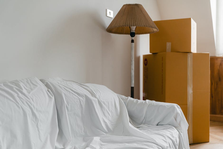 Inside a residential room showing a white fabric-covered sofa with visible creases, positioned against a plain beige wall. To the right of the sofa, there are three cardboard boxes stacked, likely use